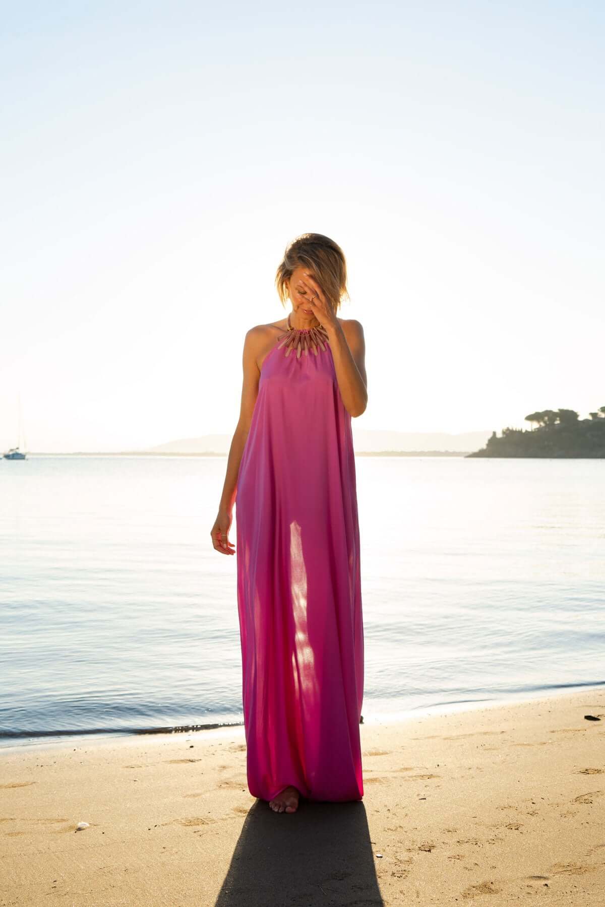 Model in a long pink silk dress standing on a beach at sunset, showcasing elegance and style.