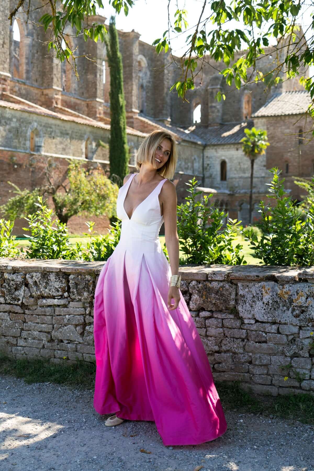 Model wearing a long cotton degradé dress with a v-neck and wide skirt, surrounded by greenery and ancient ruins.