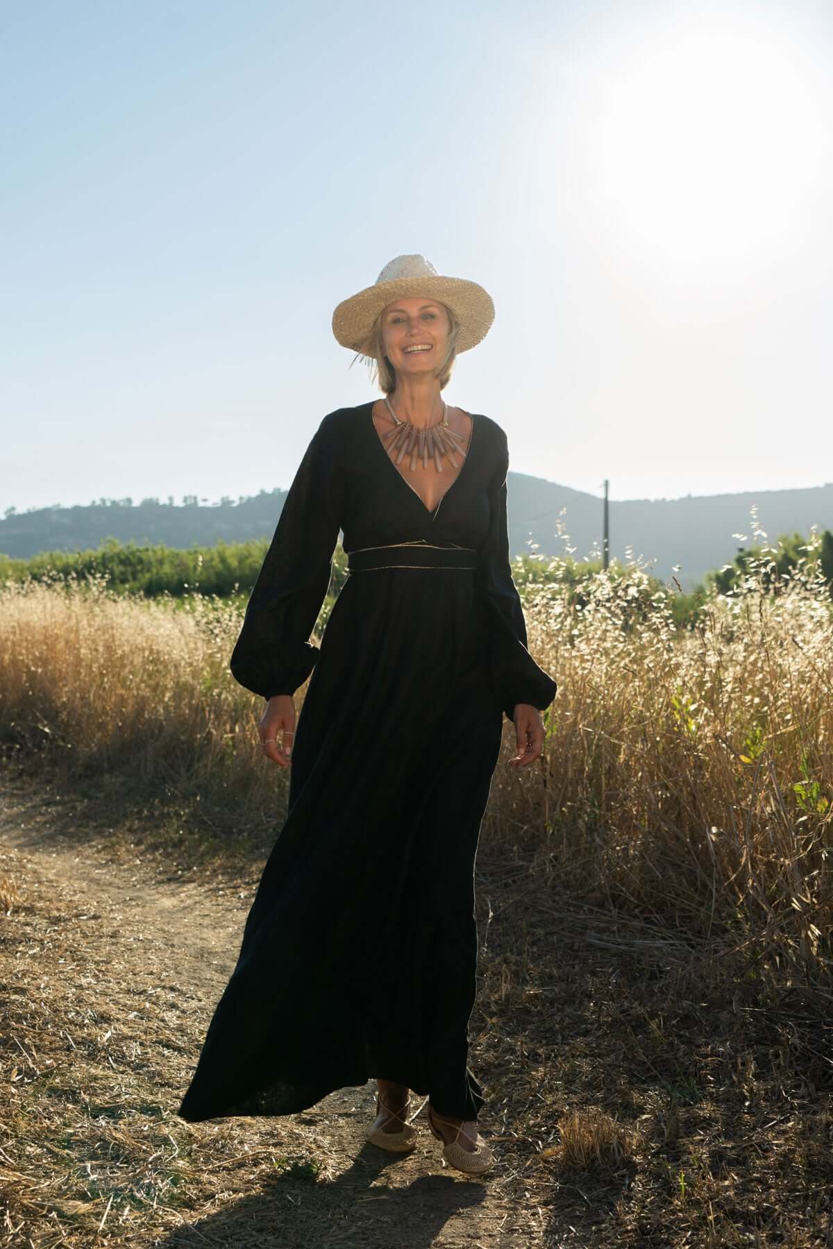 Woman in a long black dress with V-neck and puff sleeves, wearing a hat, walking in a sunny field.