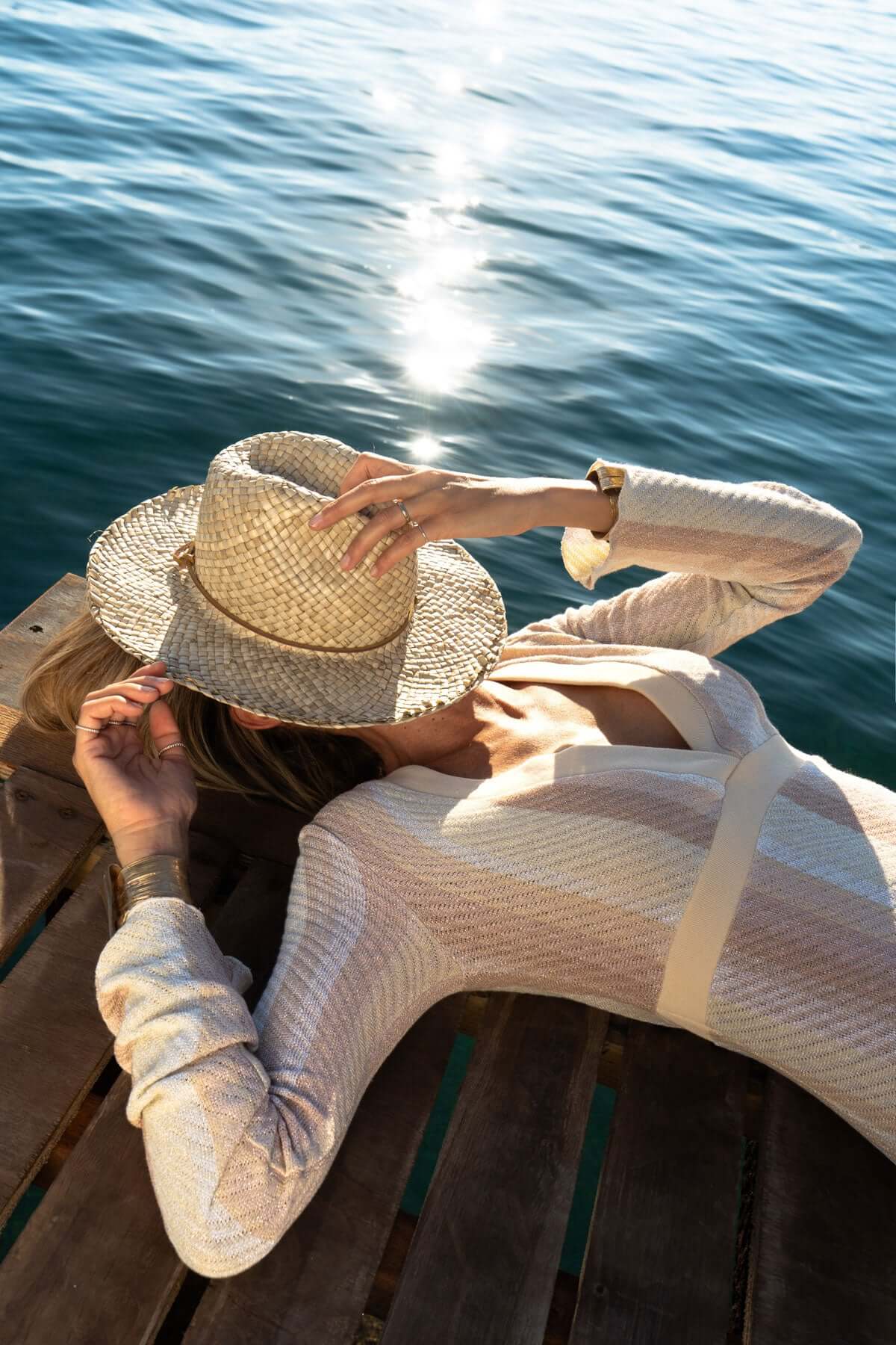 Woman in a Lurex dress and straw hat relaxing by the water on a sunny day, showcasing a serene summer vibe.